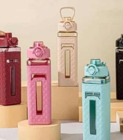 Cinq bouteilles d'eau de forme carrée disposées verticalement sur des socles cylindriques. Chaque bouteille possède un corps en plastique transparent coloré (rouge brique, rose, crème, bleu ciel et noir) partiellement recouvert d'un manchon de protection texturé assorti. Les manchons présentent un motif en relief et une fenêtre verticale transparente permettant de voir le niveau de liquide.