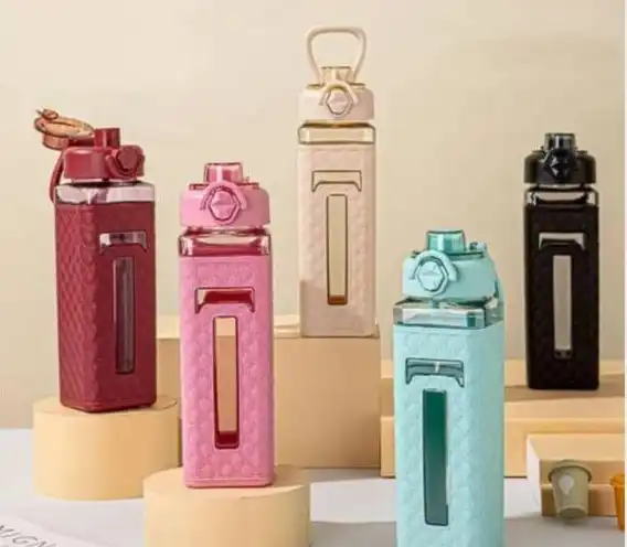 Cinq bouteilles d'eau de forme carrée disposées verticalement sur des socles cylindriques. Chaque bouteille possède un corps en plastique transparent coloré (rouge brique, rose, crème, bleu ciel et noir) partiellement recouvert d'un manchon de protection texturé assorti. Les manchons présentent un motif en relief et une fenêtre verticale transparente permettant de voir le niveau de liquide.