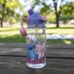 Gourde pour enfants transparente avec motif de Stitch et Angel de Disney s'embrassant avec des cœurs roses. La bouteille possède un bouchon violet avec une paille intégrée transparente déployée, un bouton-poussoir rose et une poignée souple en silicone rose, posée à l'extérieur sur une table de pique-nique en bois.