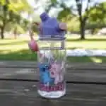 Gourde pour enfants transparente avec motif de Stitch et Angel de Disney s'embrassant avec des cœurs roses. La bouteille possède un bouchon violet avec une paille intégrée transparente déployée, un bouton-poussoir rose et une poignée souple en silicone rose, posée à l'extérieur sur une table de pique-nique en bois.