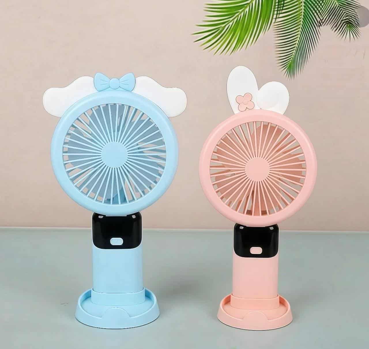 Ventilateur de Poche USB – Collection Pastel Kawaii Deux mini ventilateurs portables rechargeables au design mignon, posés côte à côte sur une surface plane vert clair. À gauche, un modèle bleu clair est décoré sur le dessus d'un nœud papillon bleu posé sur de petites ailes blanches.