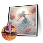 Un kit de broderie diamant montrant un tableau encadré représentant une femme de dos avec de longs cheveux clairs, vêtue d'une robe volumineuse faite de strass scintillants, entourée de grandes fleurs roses sous une pleine lune lumineuse. En bas à gauche, un gros plan circulaire met en évidence l'utilisation d'un stylet rose qui dépose précisément un petit strass brillant sur la toile quadrillée et numérotée.