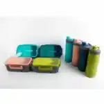Une collection de quatre kits de repas coordonnés, disposés sur un fond blanc uni. À gauche, quatre boîtes à lunch rectangulaires à compartiments sont alignées sur deux rangées ; elles possèdent des couvercles translucides colorés (bleu turquoise, vert sapin, rose corail et vert olive) avec des clips de fermeture latéraux et une cuillère intégrée visible sur le dessus