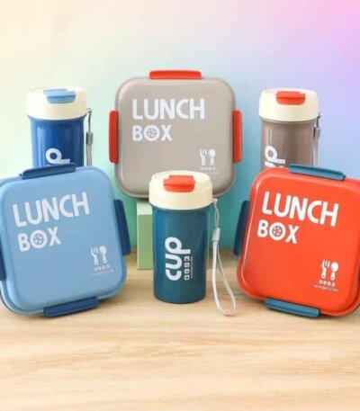 Une boîte à déjeuner carrée aux coins arrondis, avec l'inscription "LUNCH BOX" en lettres capitales blanches et une petite icône de couverts. Elle possède des clips de fermeture colorés sur les côtés.
