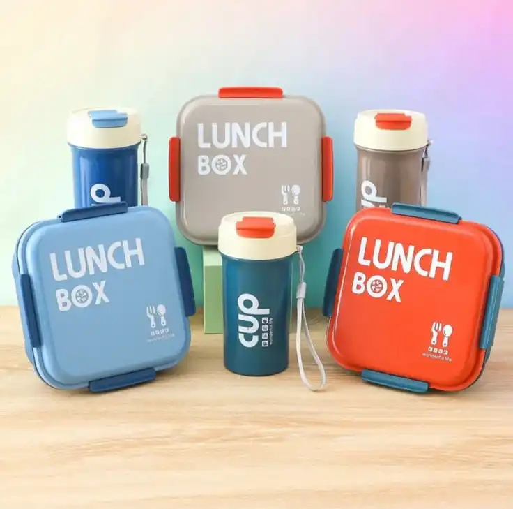 Une boîte à déjeuner carrée aux coins arrondis, avec l'inscription "LUNCH BOX" en lettres capitales blanches et une petite icône de couverts. Elle possède des clips de fermeture colorés sur les côtés.