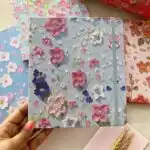 Carnet de notes rigide bleu ciel avec motifs floraux de fleurs de cerisier du Japon (sakura) blanches et roses. Le carnet dispose d'un élastique de fermeture vertical plat assorti de couleur bleu-gris sur le côté droit.