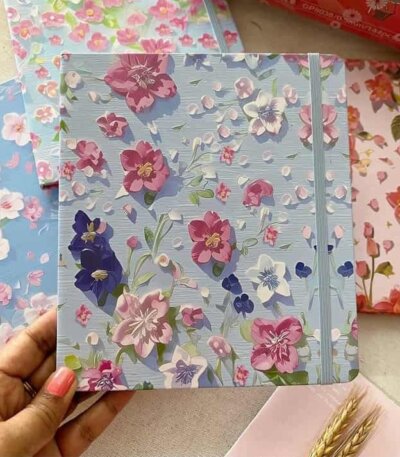 Carnet de notes rigide bleu ciel avec motifs floraux de fleurs de cerisier du Japon (sakura) blanches et roses. Le carnet dispose d'un élastique de fermeture vertical plat assorti de couleur bleu-gris sur le côté droit.