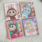Quatre carnets de notes à spirales métalliques blanches de la collection Labubu et The Monsters, disposés sur une fourrure blanche synthétique. Les couvertures en carton rigide se déclinent en rose pastel à damier, marron chocolat, rose fuchsia rétro et violet magique.