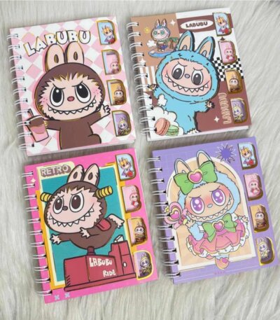 Quatre carnets de notes à spirales métalliques blanches de la collection Labubu et The Monsters, disposés sur une fourrure blanche synthétique. Les couvertures en carton rigide se déclinent en rose pastel à damier, marron chocolat, rose fuchsia rétro et violet magique.