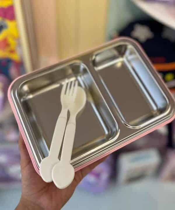 Cette photo montre une lunch box (boîte à lunch) pour enfant tenue à la main. La boîte est de forme rectangulaire avec des bords arrondis et présente une structure à deux couches : une coque extérieure en plastique rose pastel et un plateau intérieur amovible en acier inoxydable brillant.
