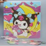 Un kit de broderie diamant représentant le personnage Kuromi de Sanrio. La toile carrée montre Kuromi avec sa coiffe noire à tête de mort rose, assise sur un croissant de lune jaune vif et tenant une petite peluche violette. L'arrière-plan est décoré d'étoiles, de cœurs roses et de vagues de couleurs pastel violettes et bleues. Toute la surface est recouverte de petits cercles numérotés prêts à recevoir les strass. Au premier plan, au pied de la toile posée à la verticale, se trouvent plusieurs sachets plastiques transparents alignés contenant les diamants colorés (rose, blanc, vert, violet).