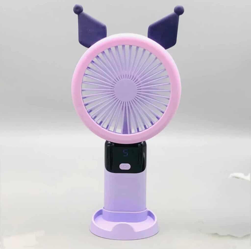 Un mini ventilateur portable rechargeable de couleur violette et mauve, inspiré du personnage Kuromi de Sanrio, posé verticalement sur son socle de table assorti. La tête ronde possède une grille rose-mauve surmontée de deux oreilles pointues de bouffon violet foncé. Le manche intègre un écran numérique noir affichant le chiffre « 5 » en bleu juste au-dessus d'un bouton d'alimentation blanc.