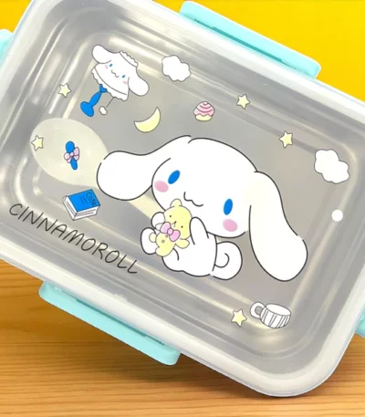 Boîte à déjeuner bento rectangulaire transparente et bleu ciel. Le couvercle transparent affiche une illustration centrale du personnage Cinnamoroll de Sanrio tenant un ourson en peluche, entouré de petites étoiles, de nuages, d'une tasse et d'un livre bleu.